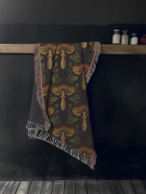 A woven cotton throw featuring an intricate mushroom and foliage pattern in deep charcoal and golden hues, framed by soft fringe.
